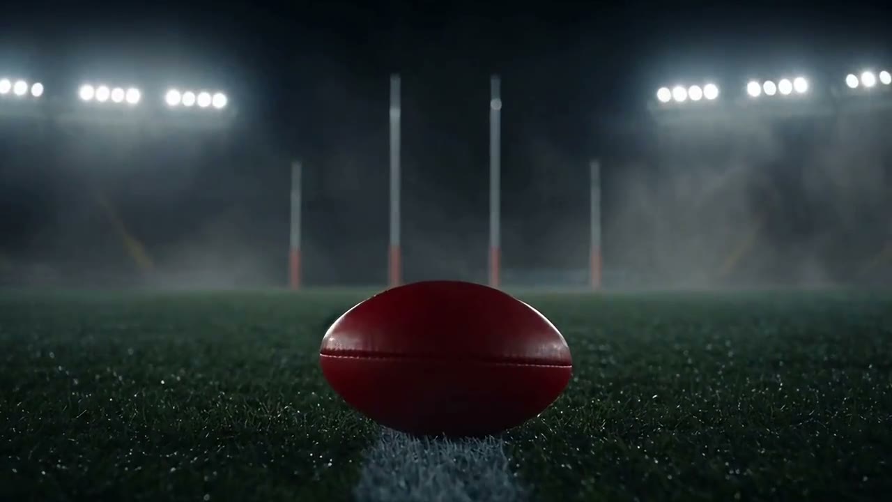 A red AFL football on the line of a wet, floodlit ground with the four goalposts behind it