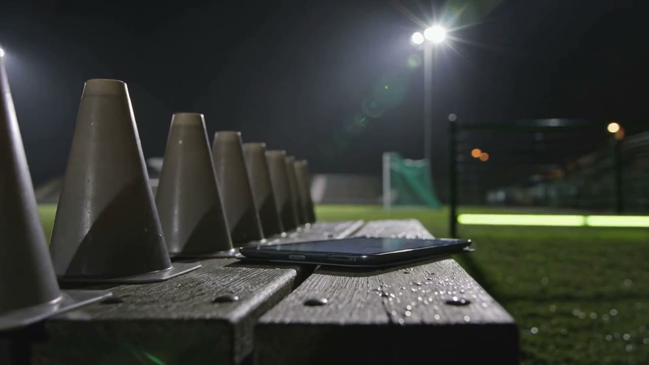 Training cones and a tablet on a wooden bench beside a floodlit community sports ground at night